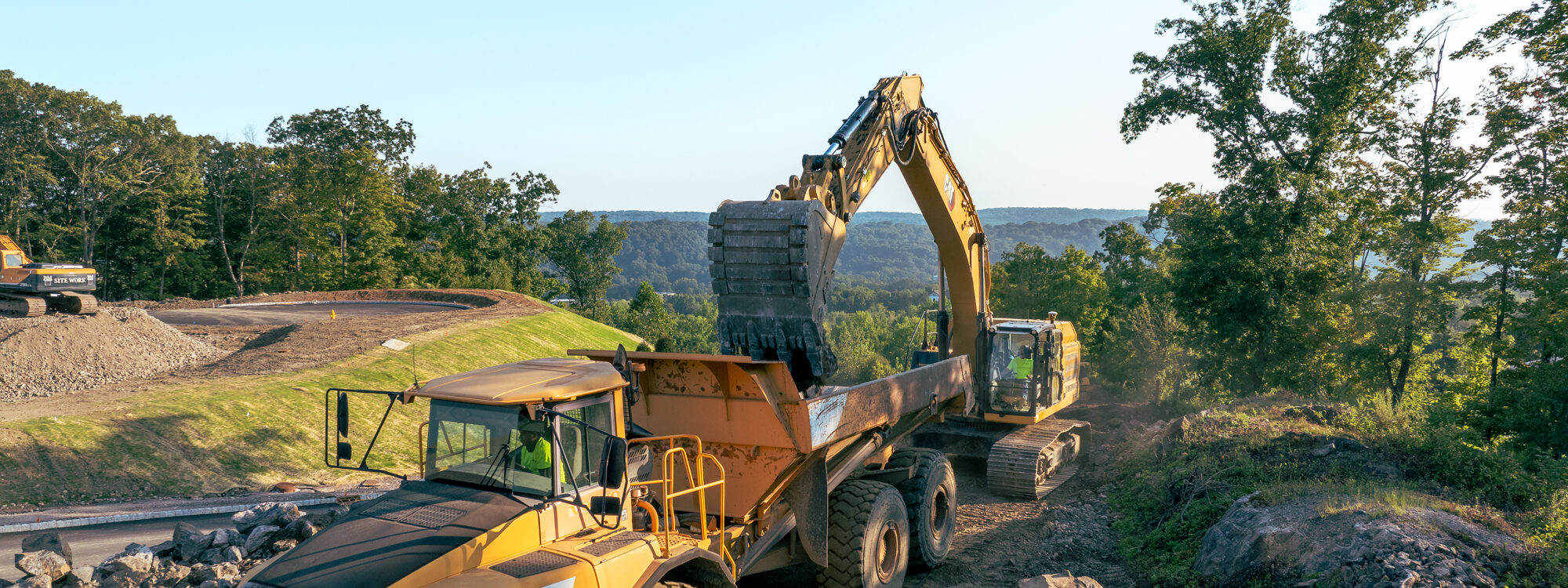 Sweeney, excavator putting dirt into haul truck, greenery and mountains in background, 24075_BUILDWITT_0771